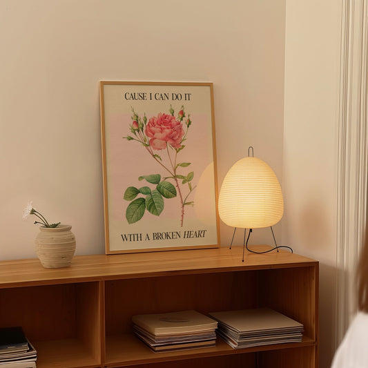 Cause I Can Do It With A Broken Heart Taylor Swift print of a coral pink rose on pale pink, framed on a shelf beside a paper lantern