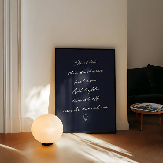 Don't Let This Darkness Fool You Noah Kahan lyric print in white handwriting on navy, framed, leaning against a wall beside a globe lamp