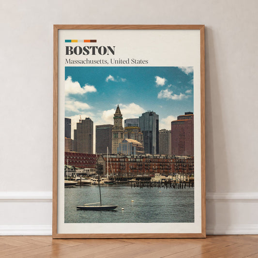 Boston travel print of the harbour skyline and Custom House clock tower behind a small sailboat, in an oak frame, leaning against a wall on a herringbone floor
