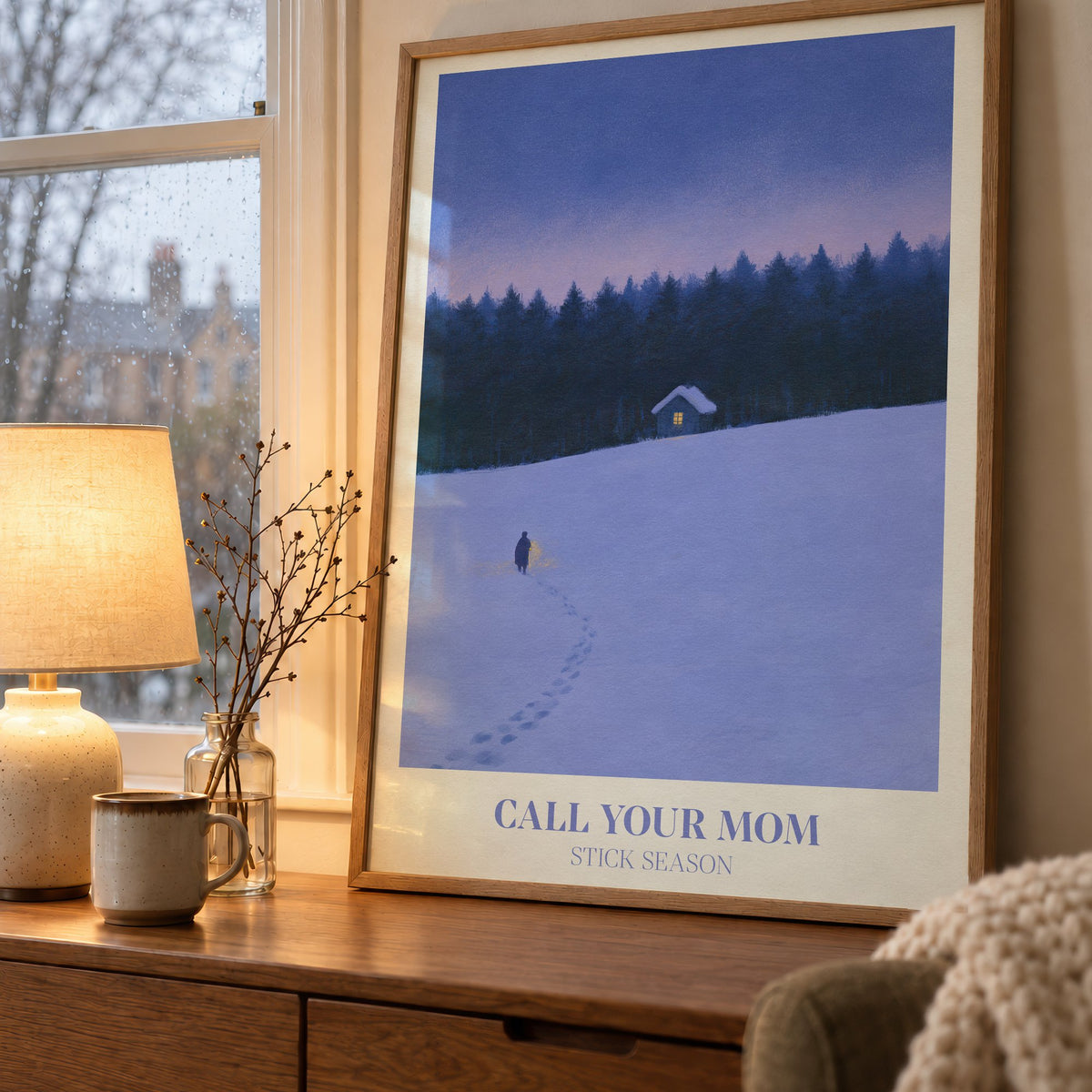Call Your Mom Winter Noah Kahan print of a lone walker heading to a lit cabin in the snow, on a wooden sideboard by a rainy window