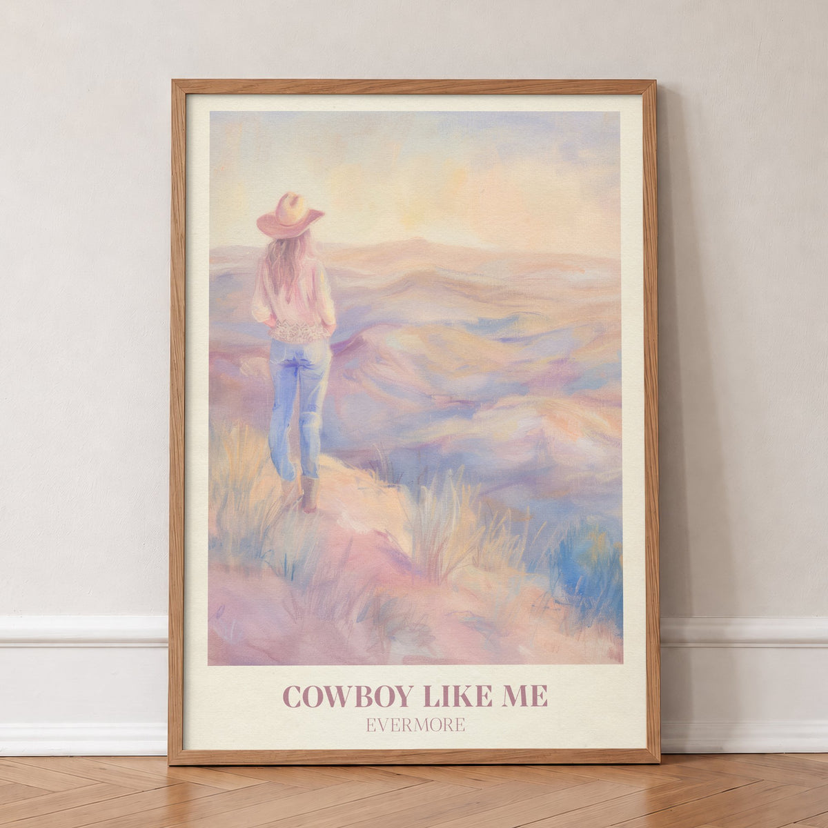 Cowboy Like Me Taylor Swift print of a cowgirl looking out over pastel hills at sunset, in an oak frame, leaning against a wall on a herringbone floor