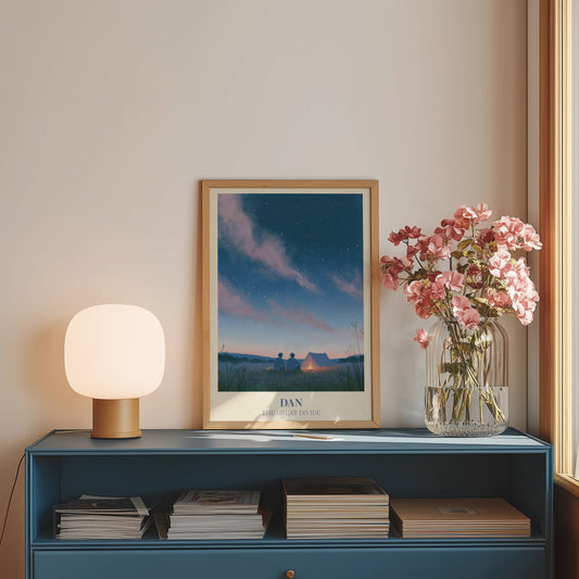 Dan Noah Kahan print of two friends by a campfire under the stars, framed on a blue sideboard beside pink flowers
