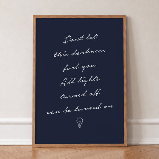 Don't Let This Darkness Fool You Noah Kahan lyric print in white handwriting on navy, in an oak frame, leaning against a wall on a herringbone floor