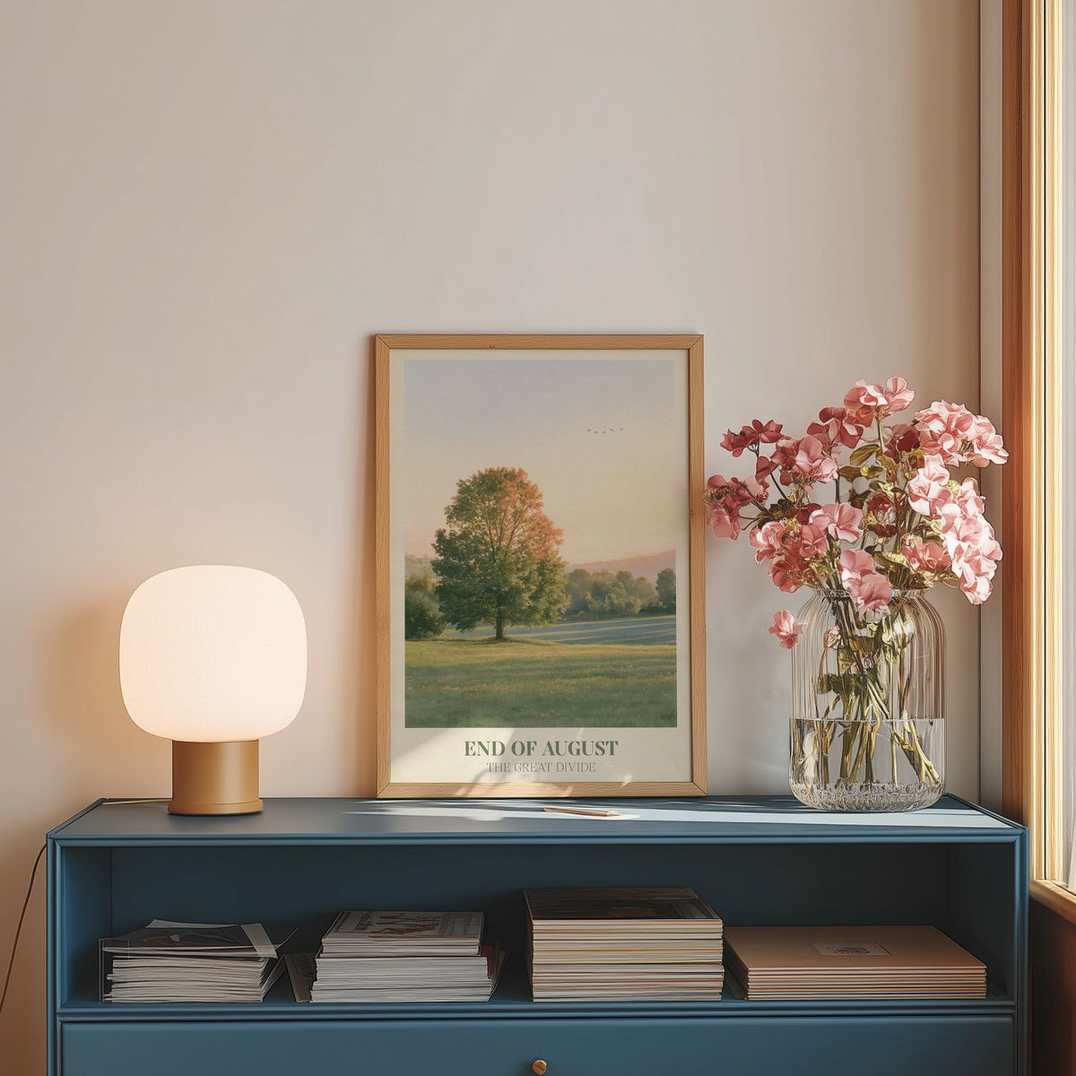 End of August Noah Kahan print of a lone tree in a late-summer field, framed on a blue sideboard beside pink flowers