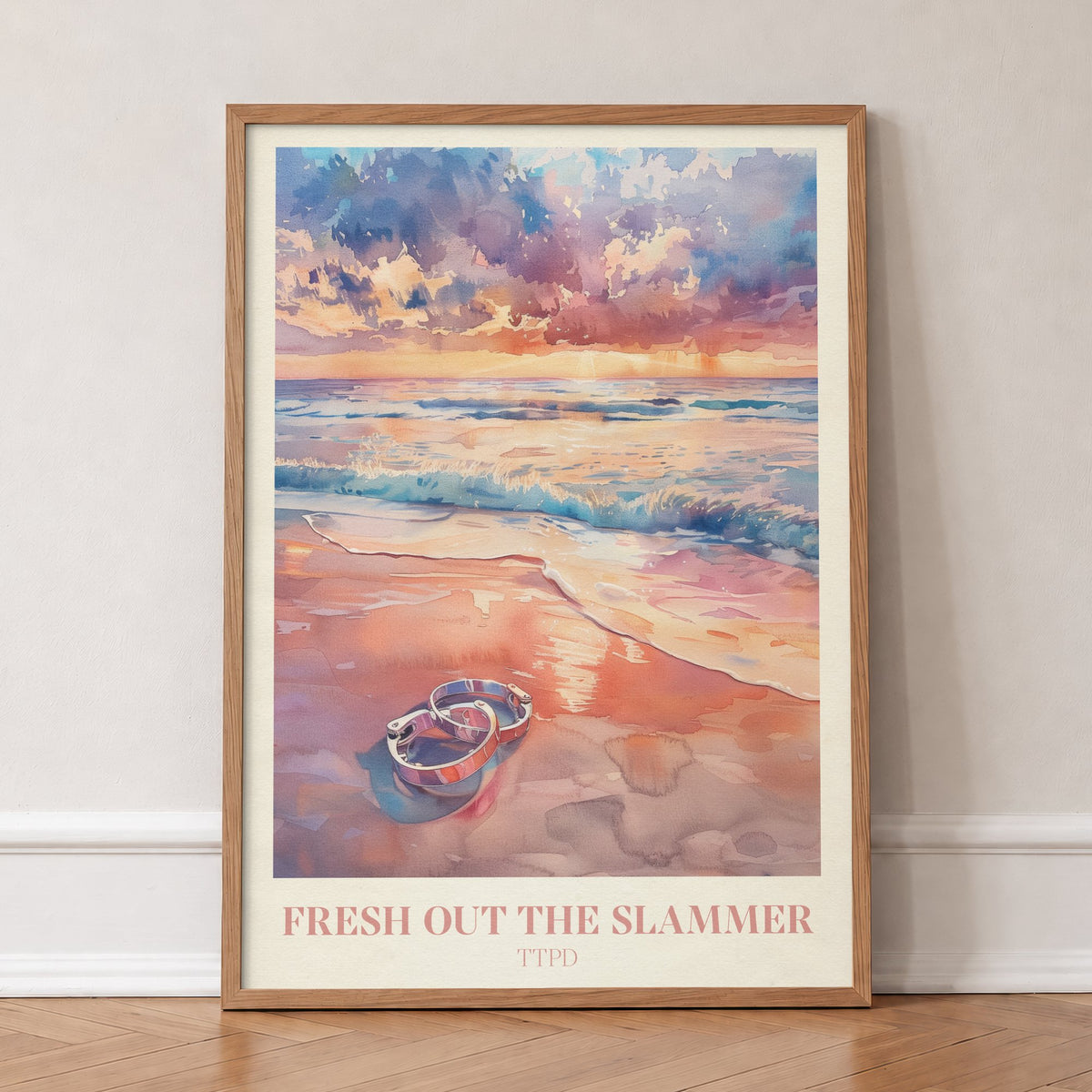 Fresh Out The Slammer Taylor Swift print of open handcuffs on wet sand at a pink sunset, in an oak frame, leaning against a wall on a herringbone floor