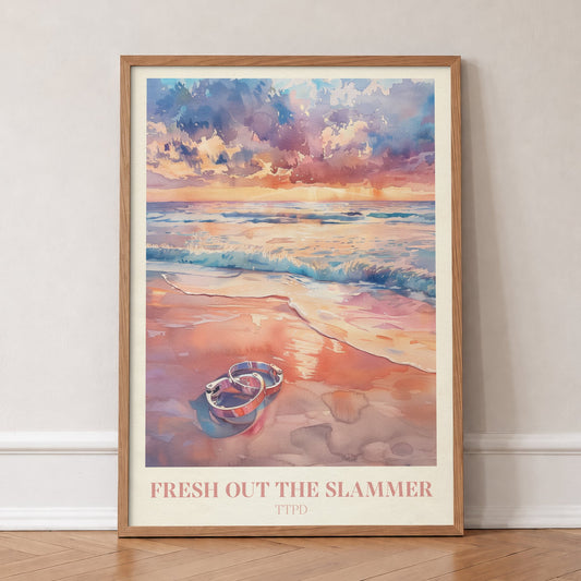 Fresh Out The Slammer Taylor Swift print of open handcuffs on wet sand at a pink sunset, in an oak frame, leaning against a wall on a herringbone floor