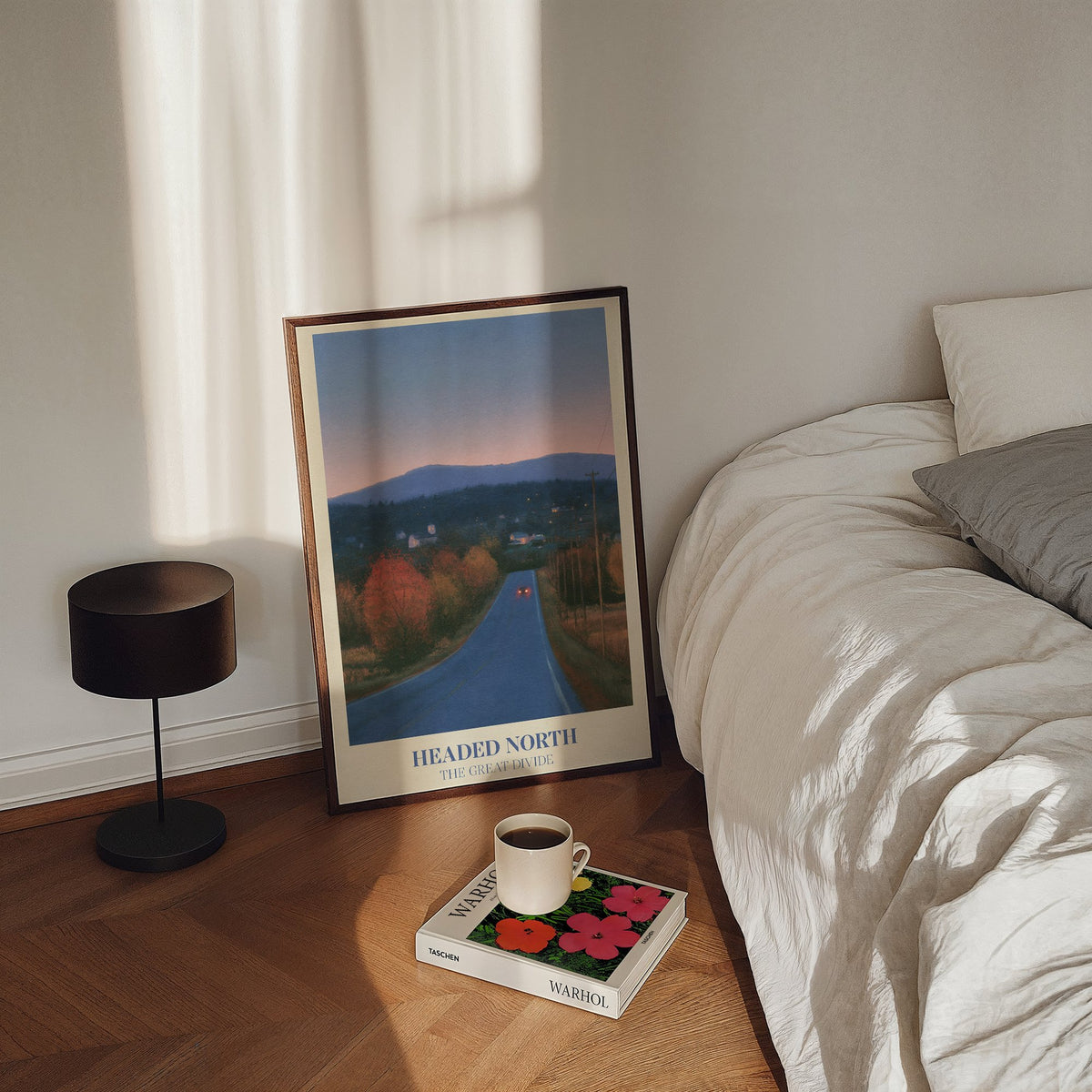 Headed North Noah Kahan print of a country road through autumn trees at dusk, framed, leaning on a bedroom floor beside a bed and a lamp