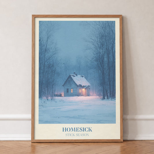 Homesick Noah Kahan print of a snowy cabin with glowing windows in a winter forest, in an oak frame, leaning against a wall on a herringbone floor