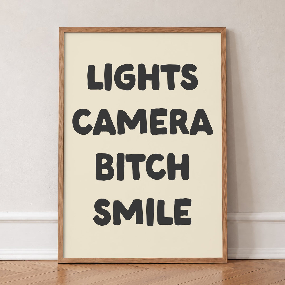 Lights Camera Bitch Smile Taylor Swift lyric print of bold black lettering on cream, in an oak frame, leaning against a wall on a herringbone floor