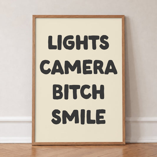 Lights Camera Bitch Smile Taylor Swift lyric print of bold black lettering on cream, in an oak frame, leaning against a wall on a herringbone floor