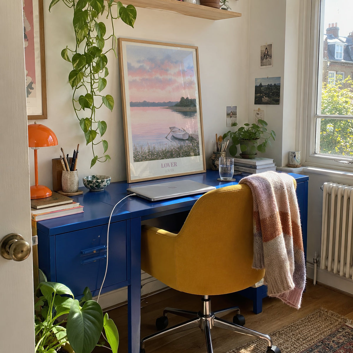 Lover Taylor Swift print of a rowing boat on a pink lake, on a blue desk in a sunny home office with trailing plants