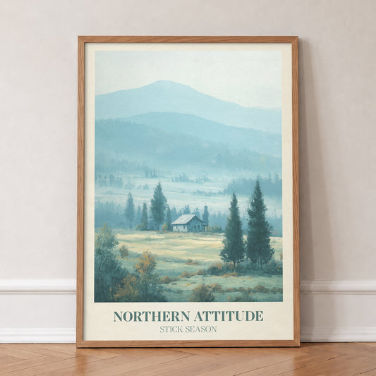 Northern Attitude Noah Kahan print of a lone cabin among pines in misty blue hills, in an oak frame, leaning against a wall on a herringbone floor