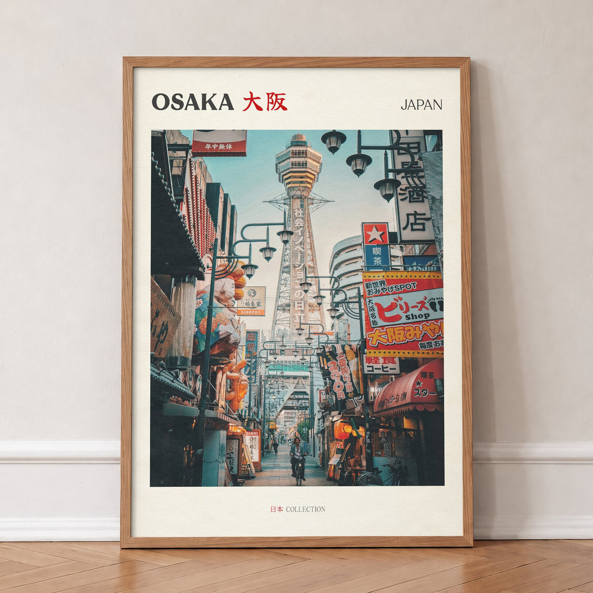 Osaka travel print of Tsutenkaku Tower rising above the neon shop signs of Shinsekai, in an oak frame, leaning against a wall on a herringbone floor