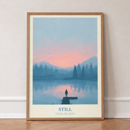 Still Noah Kahan print of a lone figure on a jetty over a pink and blue lake, in an oak frame, leaning against a wall on a herringbone floor