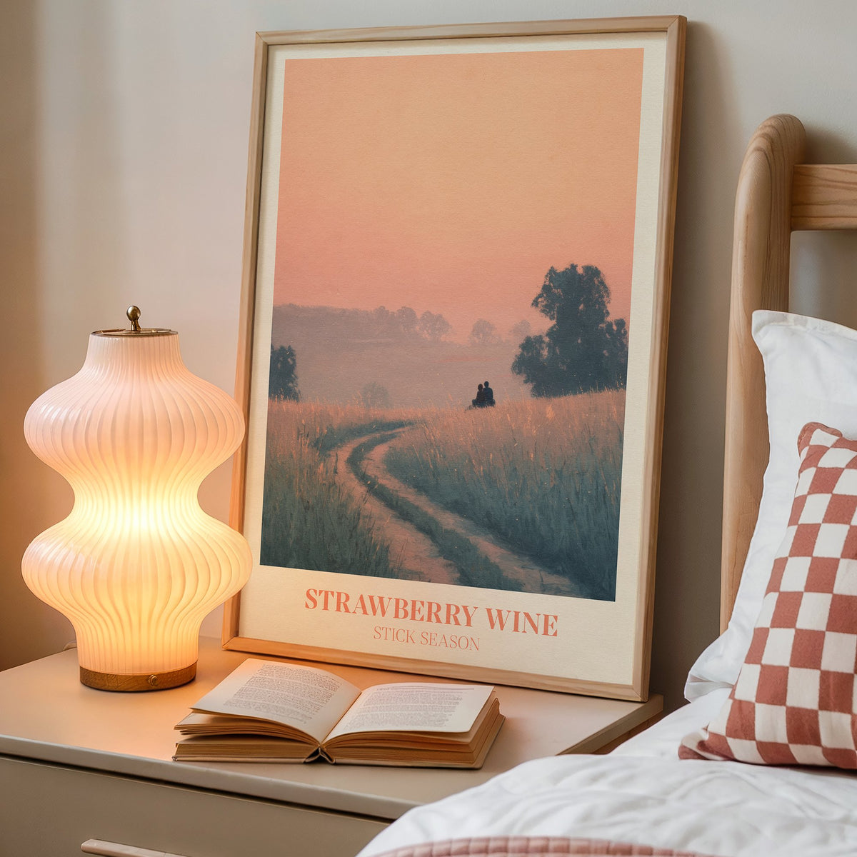Strawberry Wine Noah Kahan print of two figures in a golden meadow under a peach sky, framed on a bedside table beside a lamp