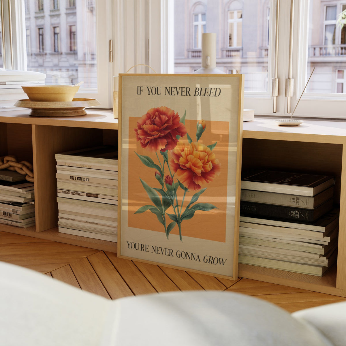 The 1 Botanical Taylor Swift print of red and orange carnations on a peach square, framed on a bookcase by a window