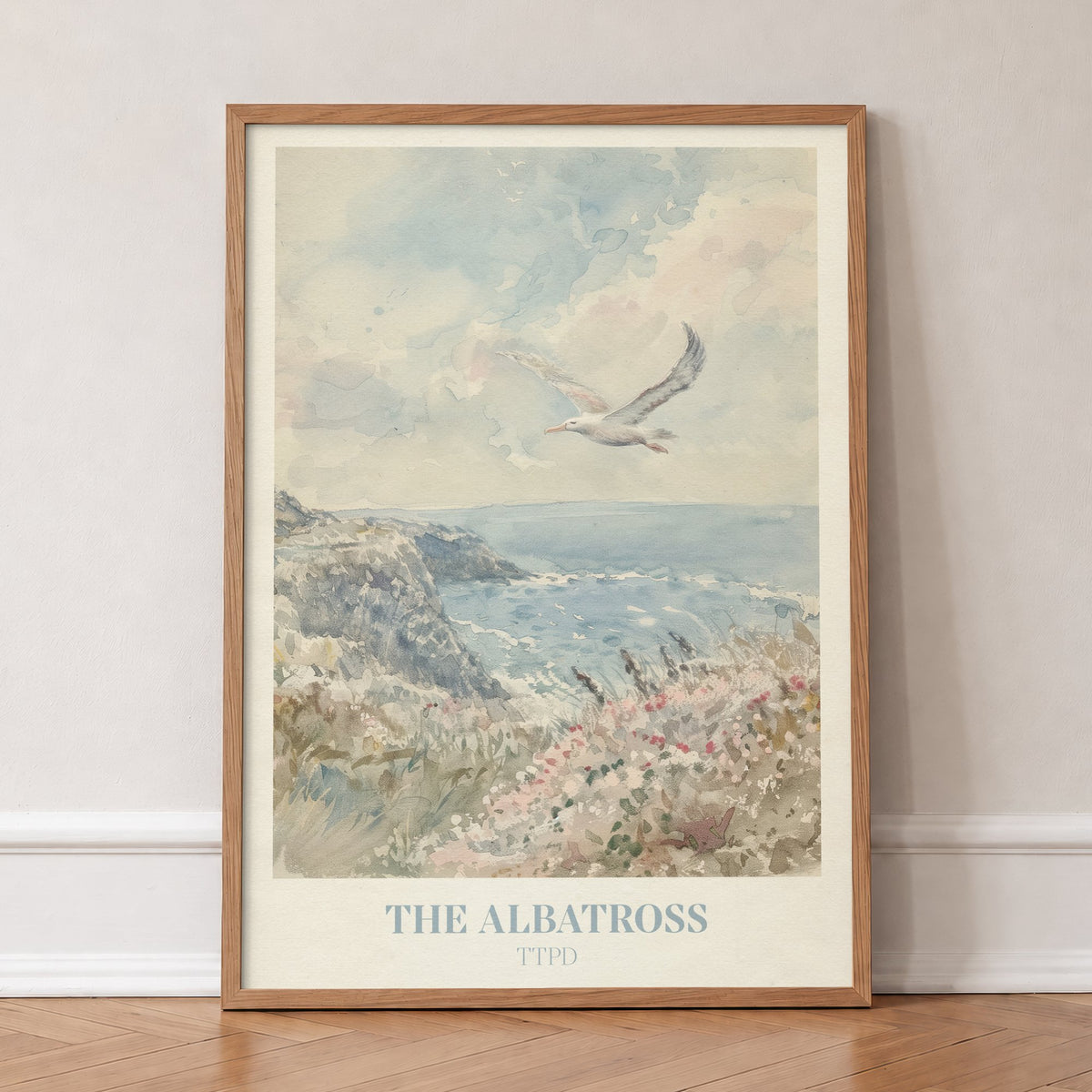 The Albatross Taylor Swift print of a seabird over a clifftop of pink wildflowers, in an oak frame, leaning against a wall on a herringbone floor