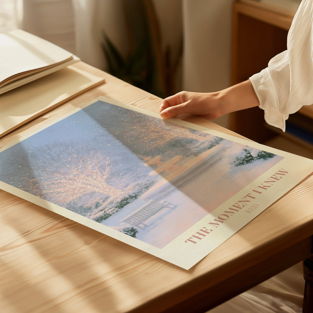 The Moment I Knew Taylor Swift print of a fairy-lit snowy park, flat on a table with a hand touching it