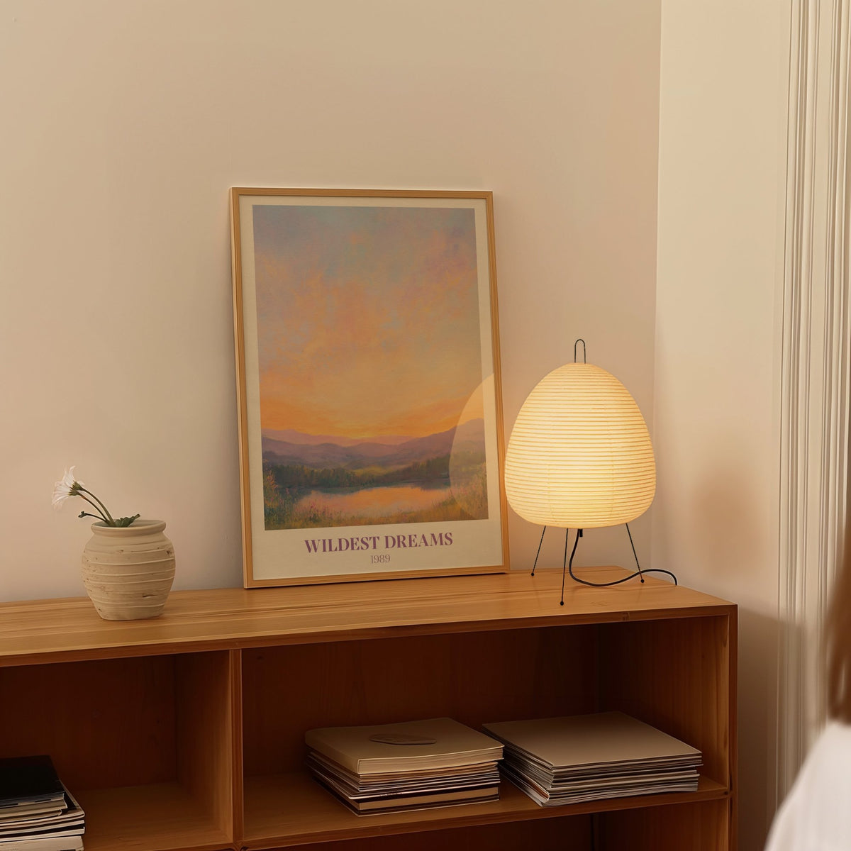 Wildest Dreams Taylor Swift print of a painted peach and lilac sunset over hills and a lake, framed on a shelf beside a paper lantern